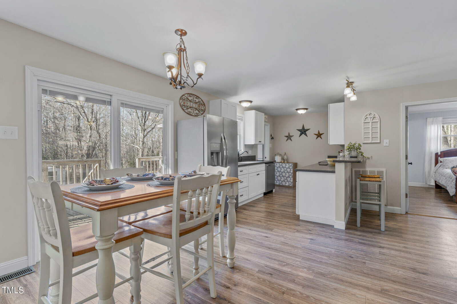 Bright kitchen and dining area with wooden floors and light-colored walls. Features a wooden table with chairs, stainless steel appliances, white cabinets, and decorative stars on the wall. Sliding doors lead to an outdoor deck with trees visible.