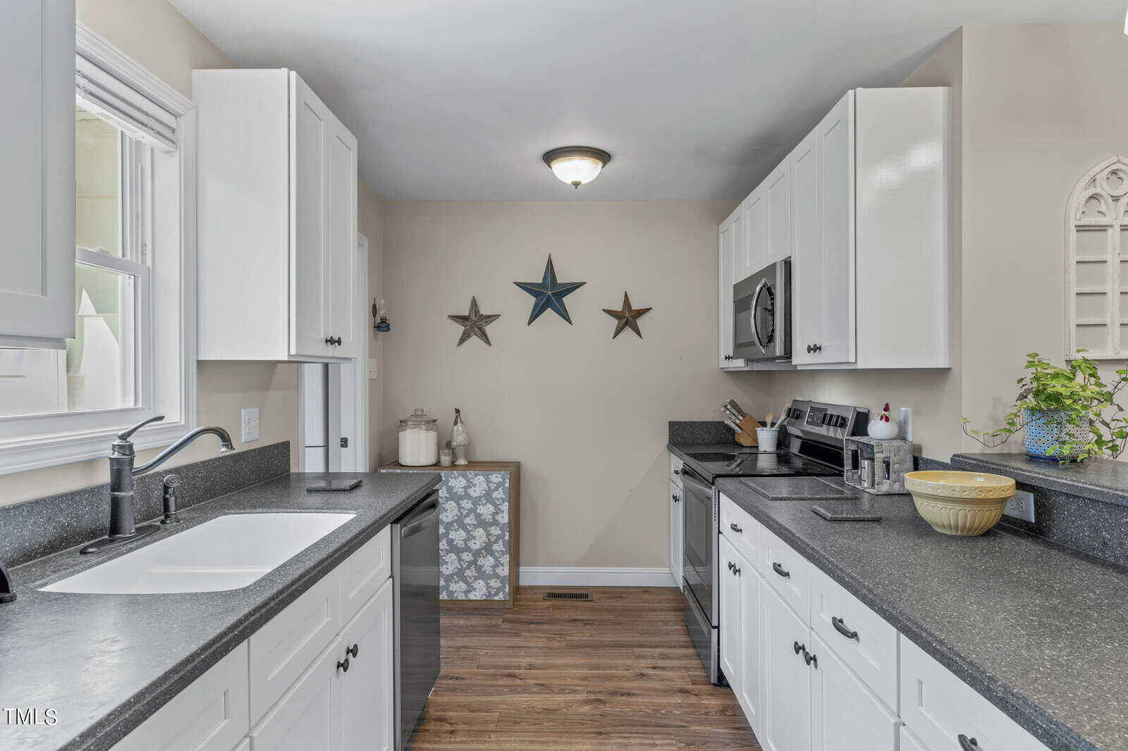 A modern kitchen with white cabinets and dark countertops features stainless steel appliances, including an oven and microwave. Three star-shaped wall decorations hang above a storage bench. A potted plant sits on the counter near the stove.