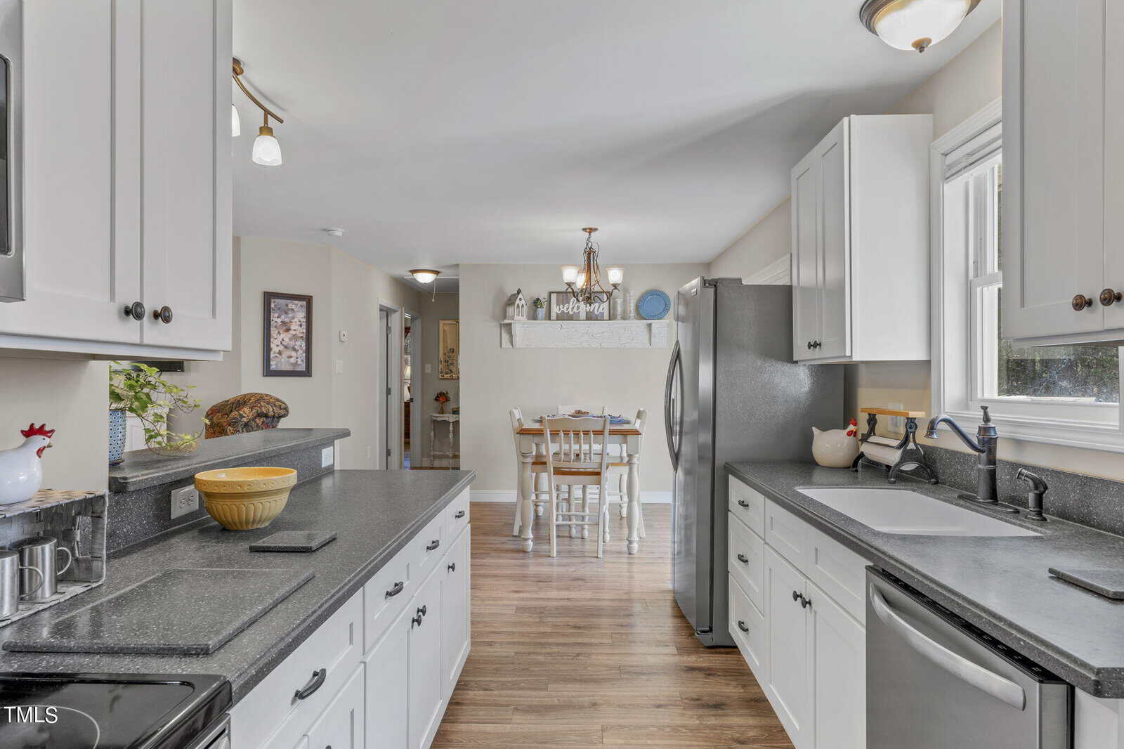 A bright kitchen with white cabinets and dark countertops. Stainless steel appliances are present, including a refrigerator and dishwasher. A dining area with a wooden table and chairs is visible in the background. The floor is hardwood.