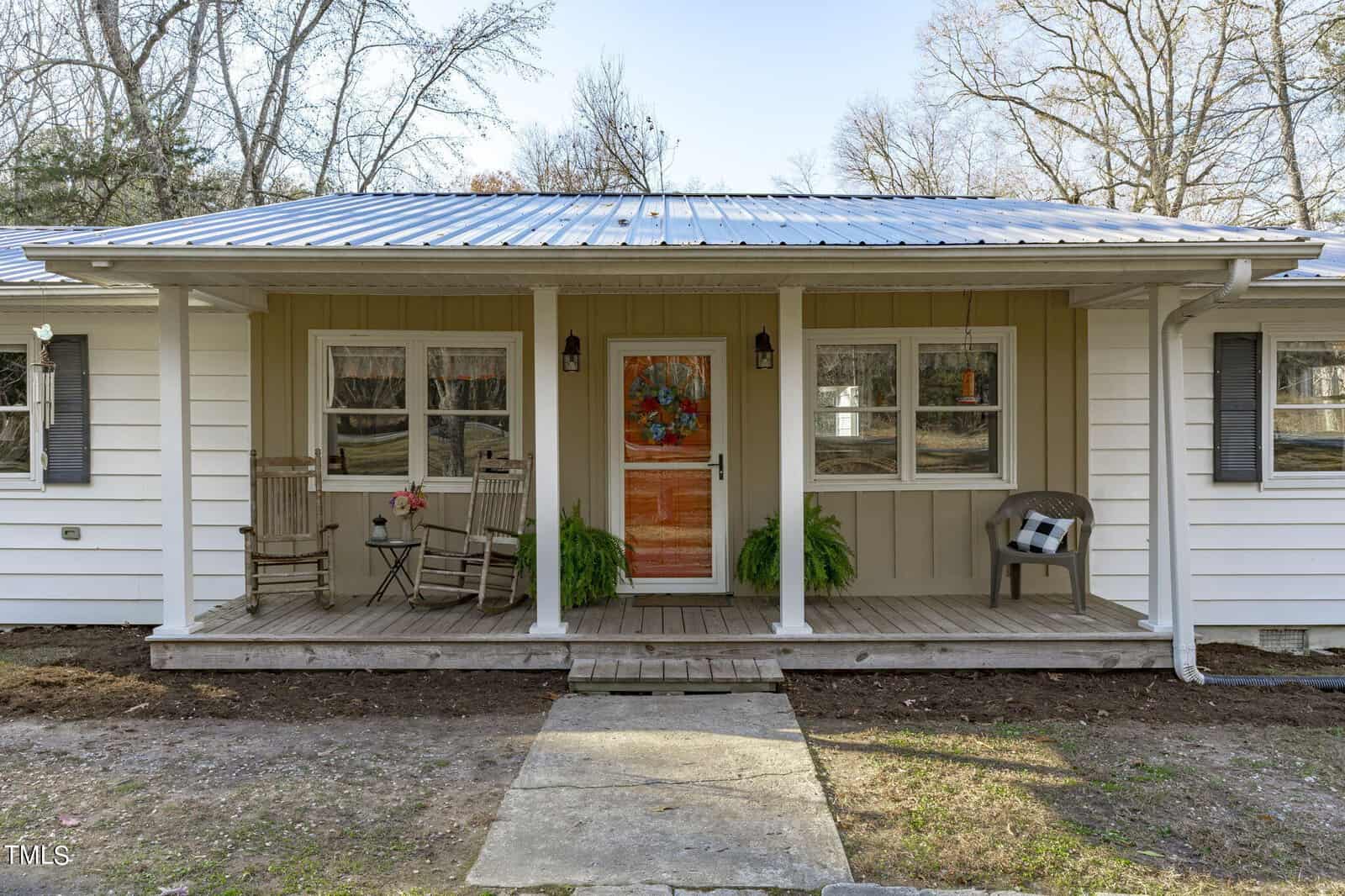 A cozy white house at 2611 Tar River Road in Creedmoor boasts a front porch with two rocking chairs and a small table on the left, and a bench draped with a blanket on the right. Adorned with plants and a door wreath, its charm is enhanced by the metal roof and tree-lined backdrop.