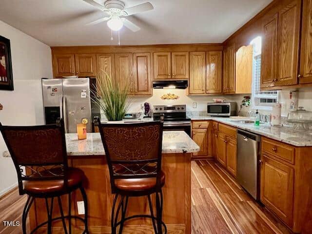 A well-lit kitchen featuring wooden cabinets, stainless steel appliances, hardwood floors, and a kitchen island with two bar stools.
