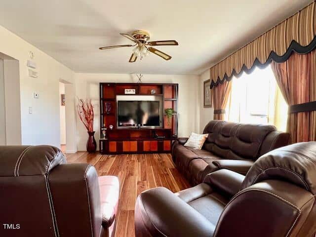 A living room featuring a brown leather sofa and loveseat, hardwood floors, a television in a wooden entertainment center, and windows with patterned curtains. a ceiling fan with lights is mounted above.