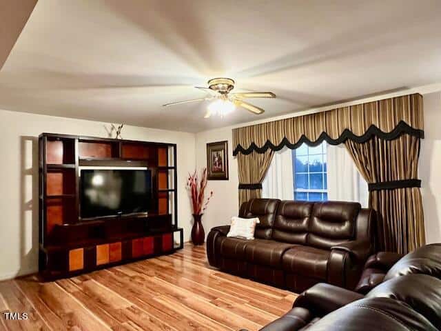 A living room featuring a brown leather sofa set, a wooden entertainment center with a mounted television, hardwood floors, a ceiling fan with lighting, and windows dressed with patterned curtains.