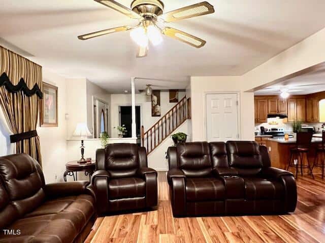 A well-lit living room with hardwood floors, featuring brown leather sofas, a ceiling fan above, and an open plan view into the kitchen.