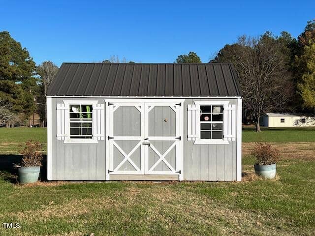 A gray garden shed with white trim and a gabled roof is situated on a grassy area with plants in containers on either side of the doors, under a clear blue sky.