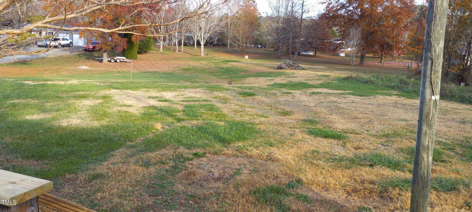 A grassy yard with patches of green and brown grass, bordered by leafless trees with orange and red leaves. A trailer is visible in the background, and the ground has a few scattered branches. The scene suggests a late autumn setting.