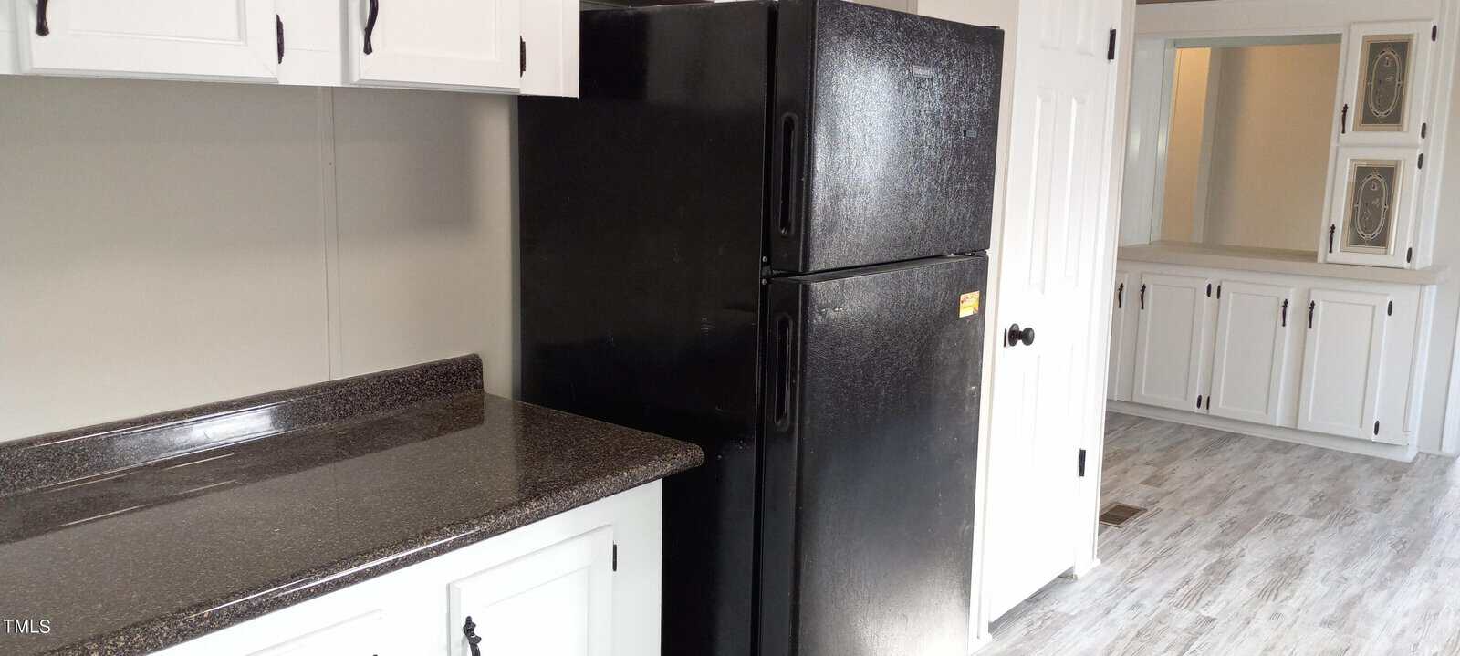 A kitchen with white cabinets and a black refrigerator. The countertop is dark speckled and contrasts with the light wood flooring. In the background is a white built-in cabinet with glass doors.