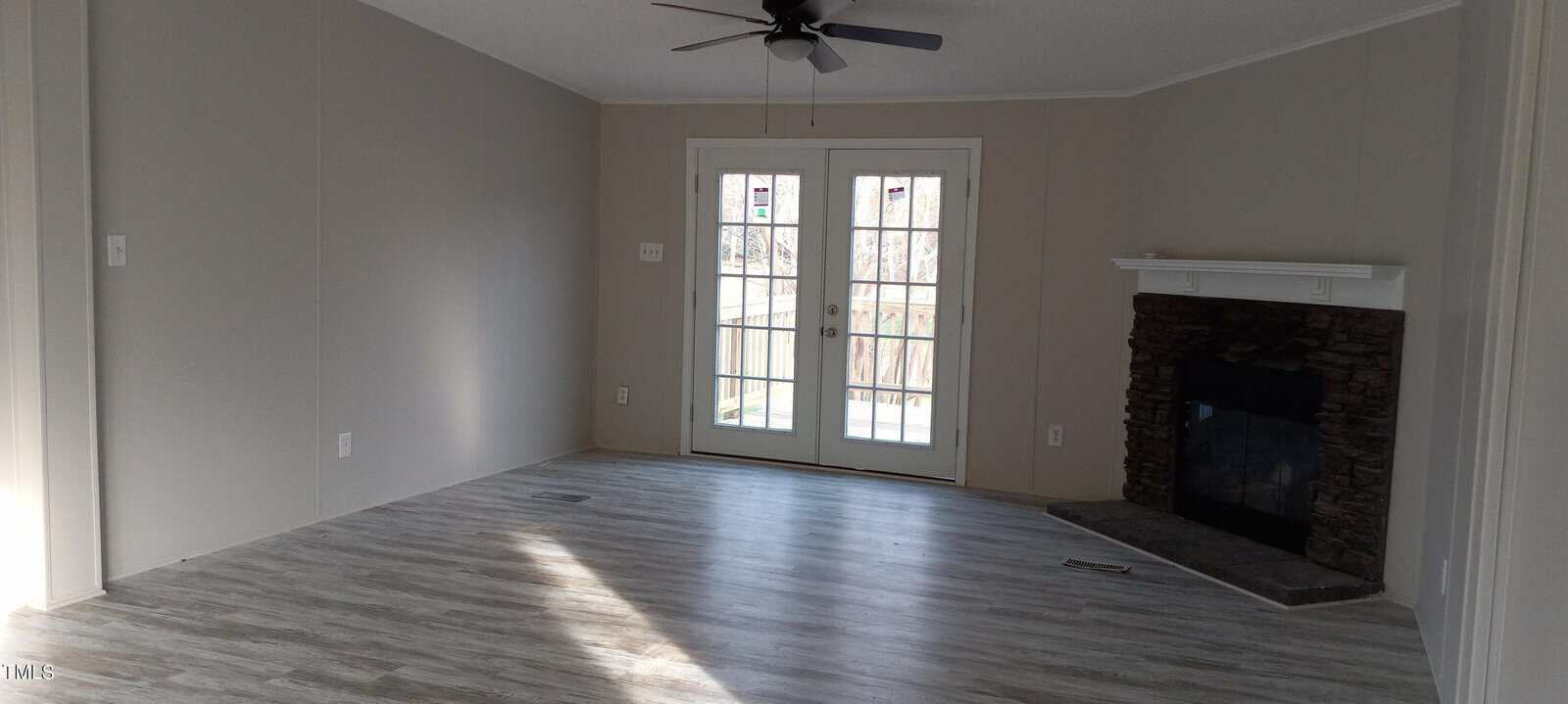 A bright, empty room with gray wood flooring features a ceiling fan and a stone fireplace on the right. Double glass doors open to a balcony, letting in ample natural light. The walls are painted light gray.