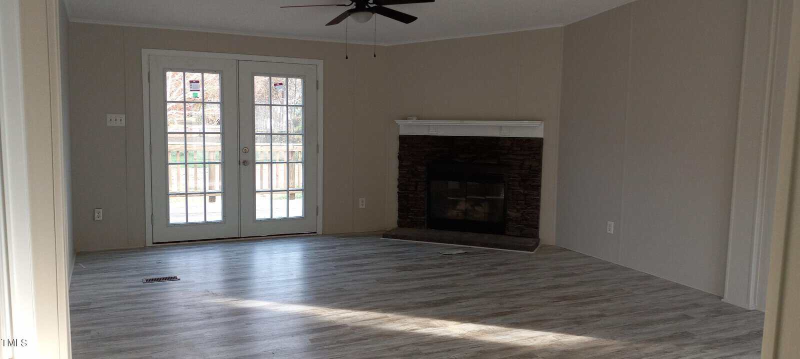 A bright living room with light wood floors, a stone fireplace, and double glass doors leading outside. A ceiling fan is mounted on the white ceiling, and natural light floods the space through the doors and a window.