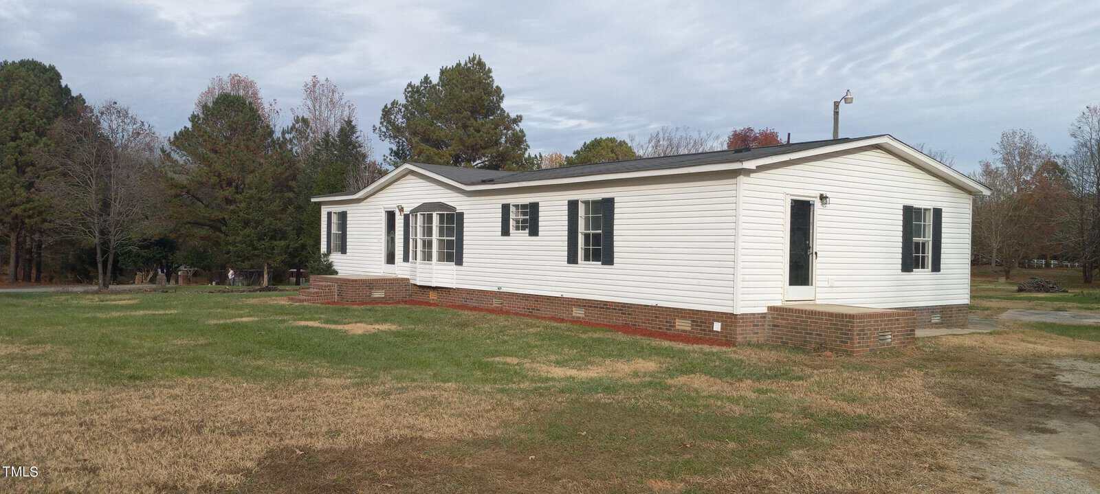 Located at 4142 Deer Run Drive in Oxford, this cozy single-story white mobile home rests on a brick foundation amidst a grassy lot. Featuring multiple windows and a charming front porch, it is beautifully framed by trees under a partly cloudy sky.