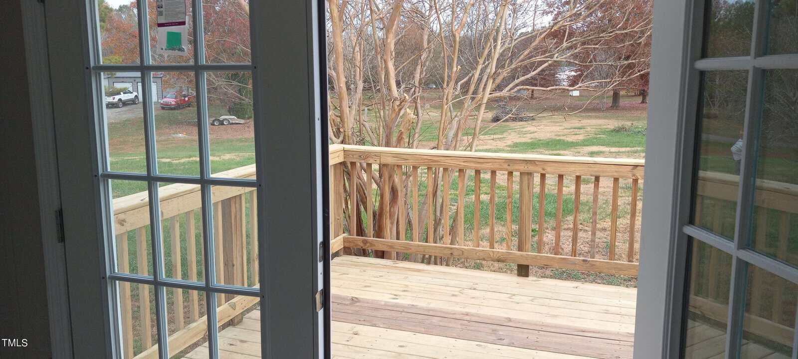 View from an open glass door leading to a wooden deck. The deck overlooks a grassy yard with bare trees and a parked vehicle visible in the distance, under a cloudy sky.