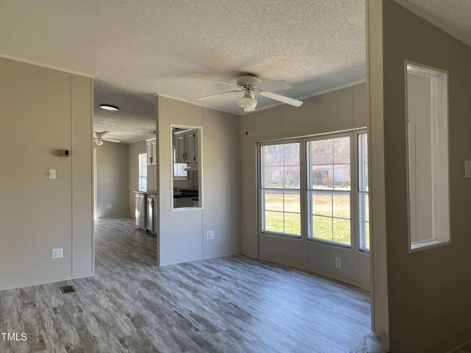 A bright, empty room with light wood flooring and large windows overlooking a yard. The room has neutral-colored walls, a ceiling fan, and opens into a kitchen area with visible countertops and appliances.