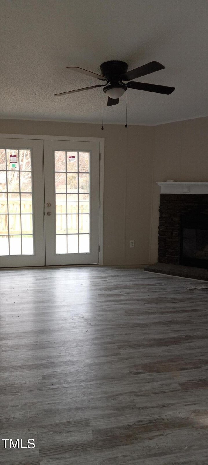 A room with light wood flooring and beige walls features a ceiling fan and a stone fireplace on the right. French doors on the left open to a fenced backyard, letting in natural light.