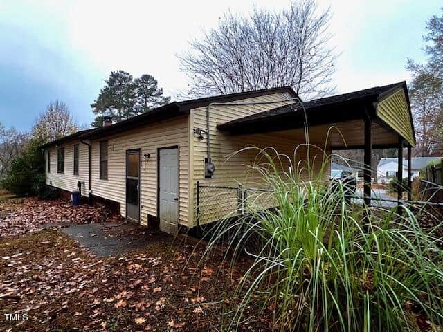 A one-story beige house with a dark roof and an attached carport on a cloudy day, surrounded by bare trees and overgrown grass.