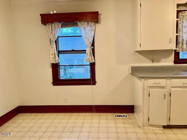 A bare kitchen interior with a single window adorned with curtains, an empty countertop with a sink, and a tiled floor.