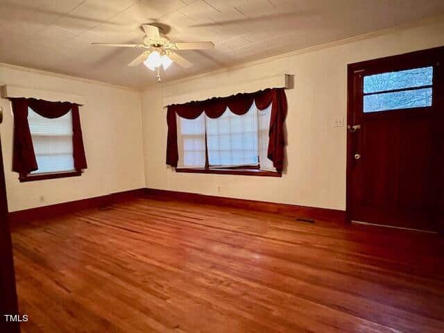 An empty room with hardwood floors, a ceiling fan, and two windows with dark curtains. there is a red door partially visible on the right.