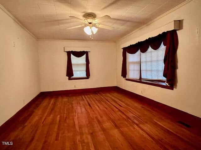 An empty room with wooden flooring, a ceiling fan, and two windows with red valance curtains.