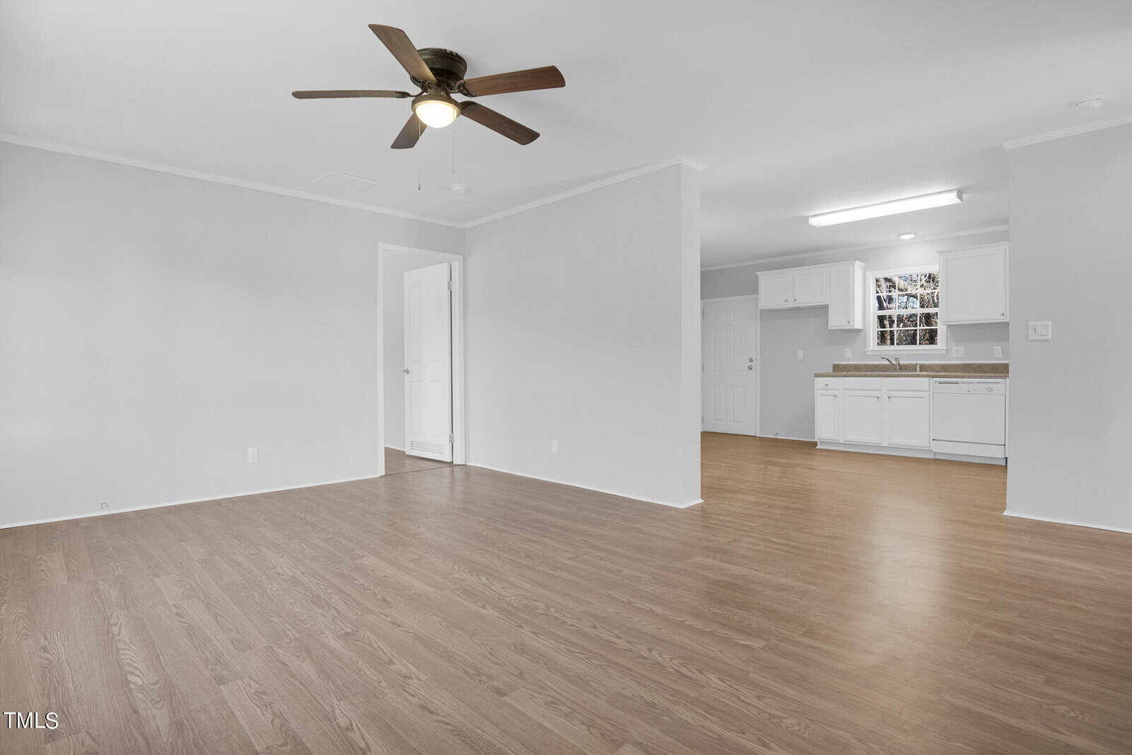 An empty, spacious room with light hardwood flooring, white walls, and a ceiling fan. the room opens to a white kitchen with cabinets and a small dining area. a doorway leads to another room.