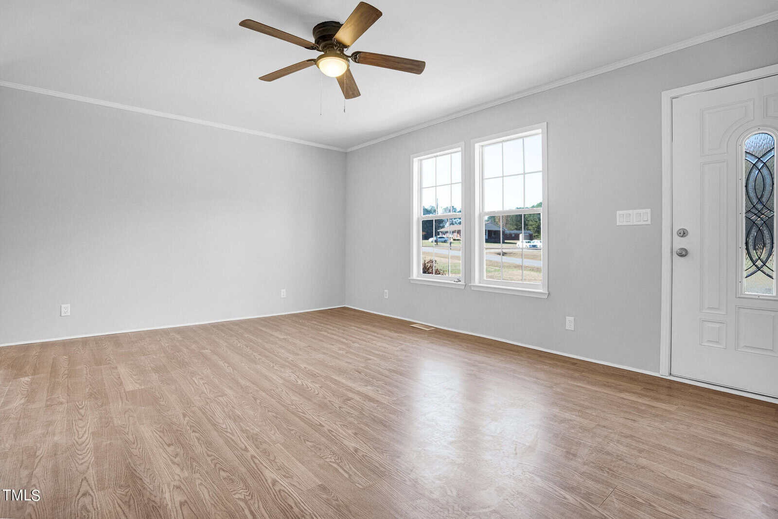 Empty living room with gray walls and light wooden flooring. the room features a ceiling fan, a front door with decorative glass, and two large windows overlooking a street.