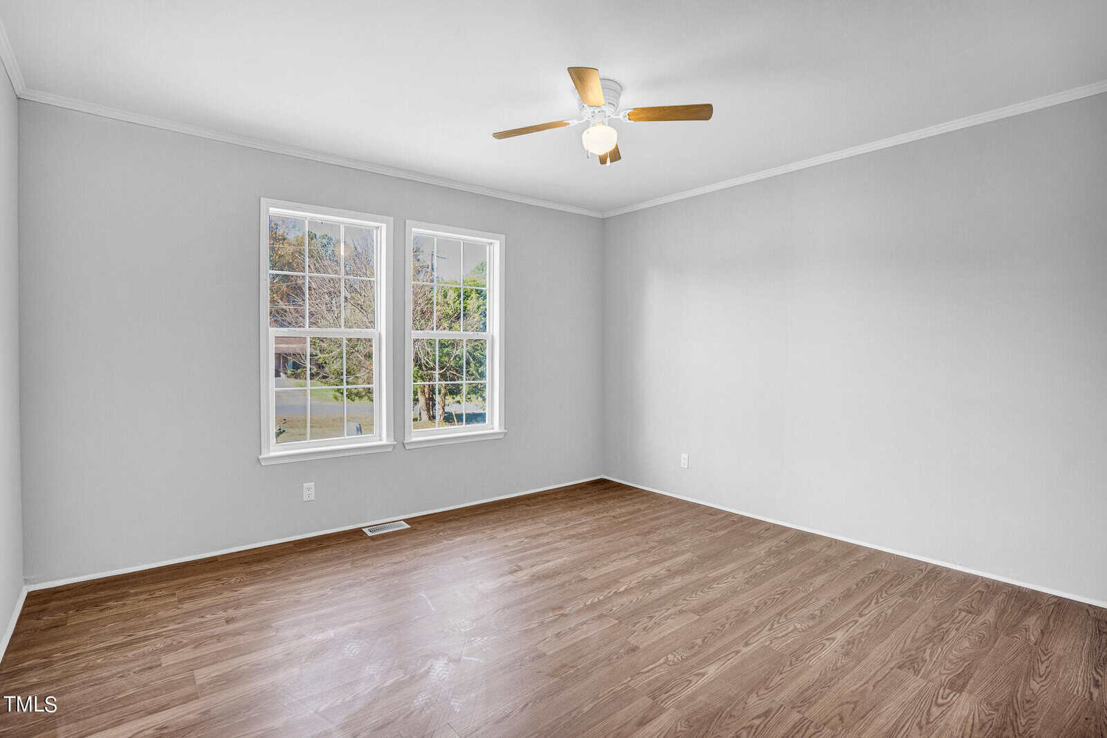 Empty room with white walls, brown wooden flooring, and a ceiling fan with light. two large windows provide a view of the outside.