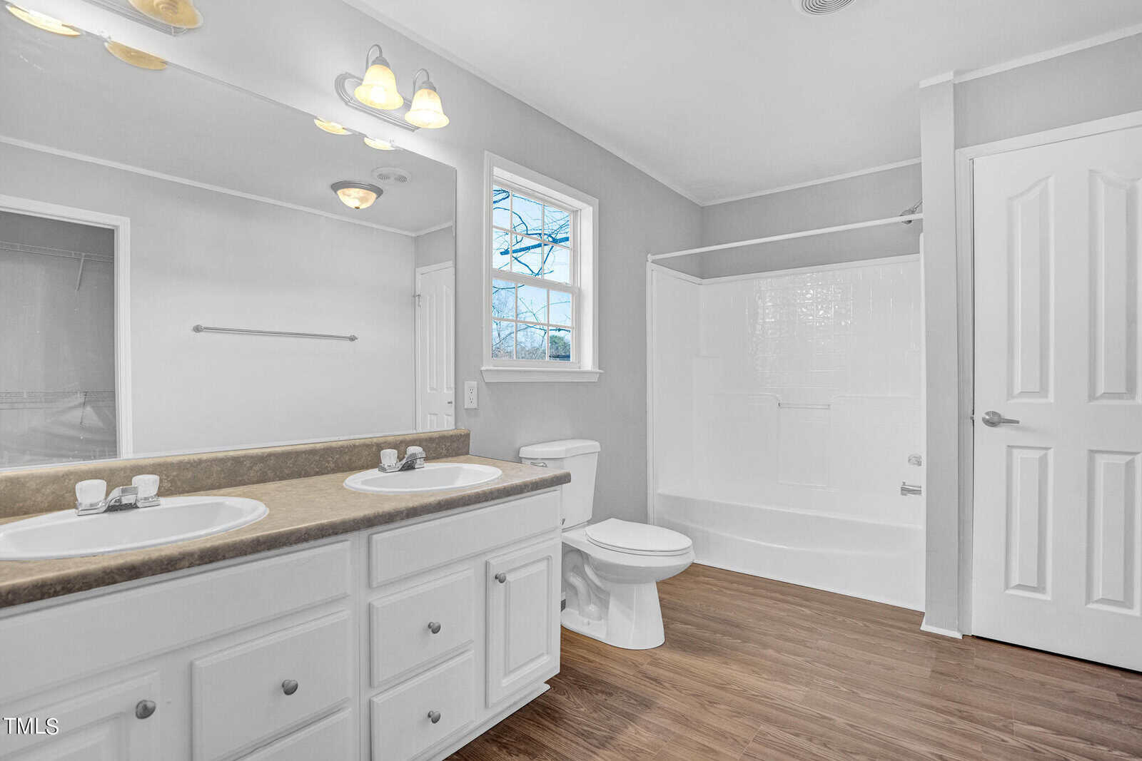 A bright bathroom featuring white cabinetry with dual sinks, a large mirror, a bathtub-shower combo, a toilet, and a window providing natural light. the flooring is wooden, and the walls are painted white.