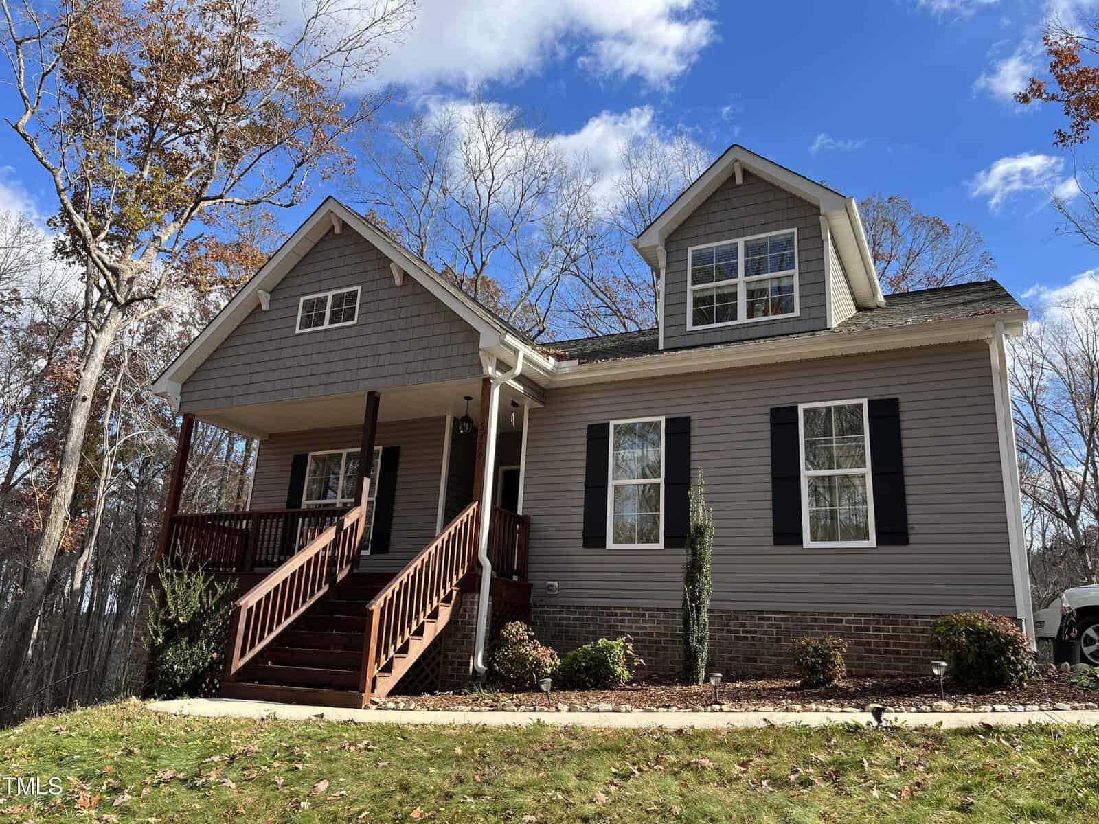 Nestled at 2480 Golden Forest Drive in Franklinton, this charming gray house features a front porch with wooden steps and a gabled roof. It's set against tall trees with autumn foliage, complementing the symmetrical windows beneath a partly cloudy sky.