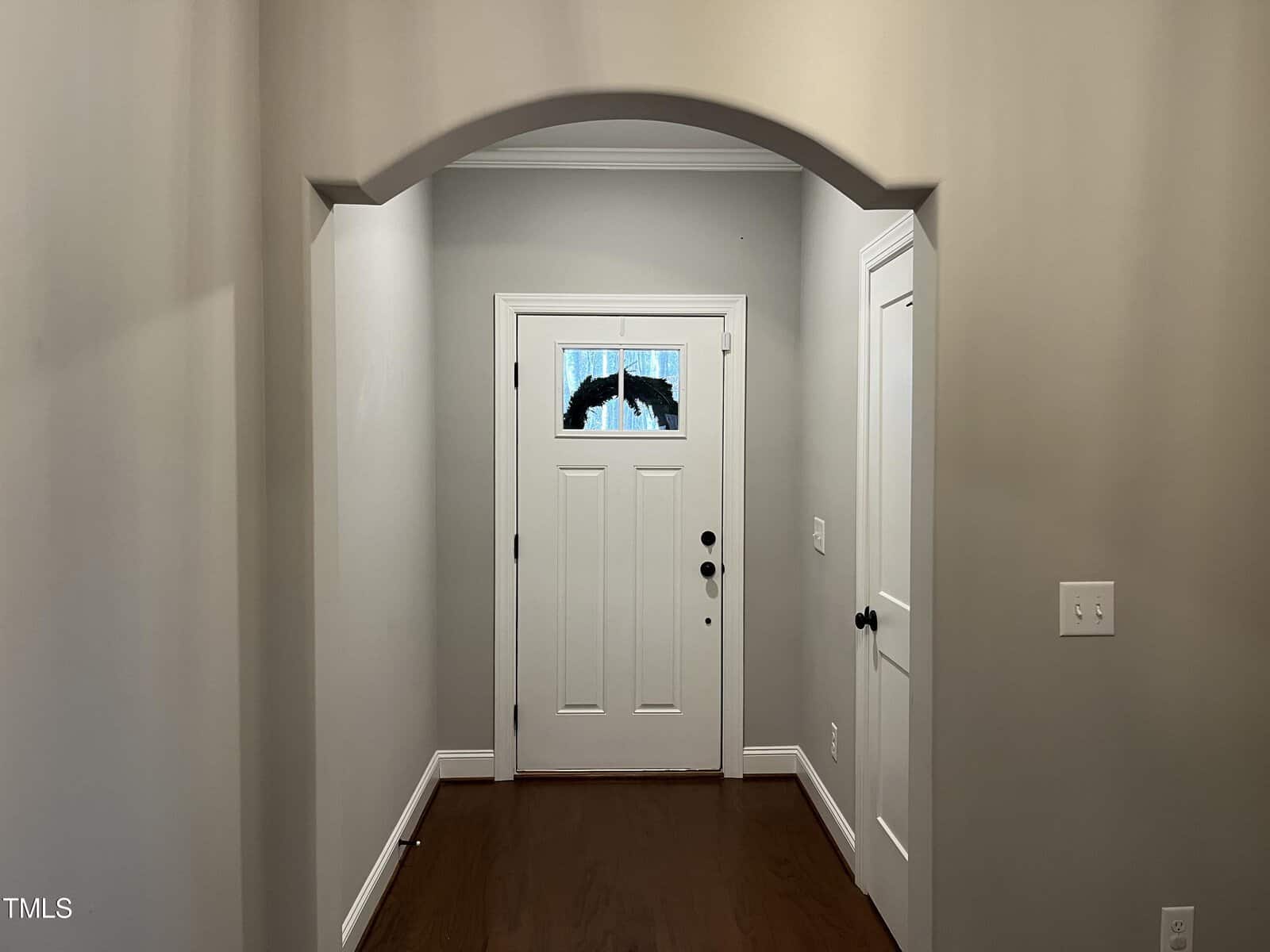 A hallway with light gray walls leading to a white front door with an arched design above. The door has a small window with a decorative piece. Two additional white doors are on the sides. The floor is dark wood.