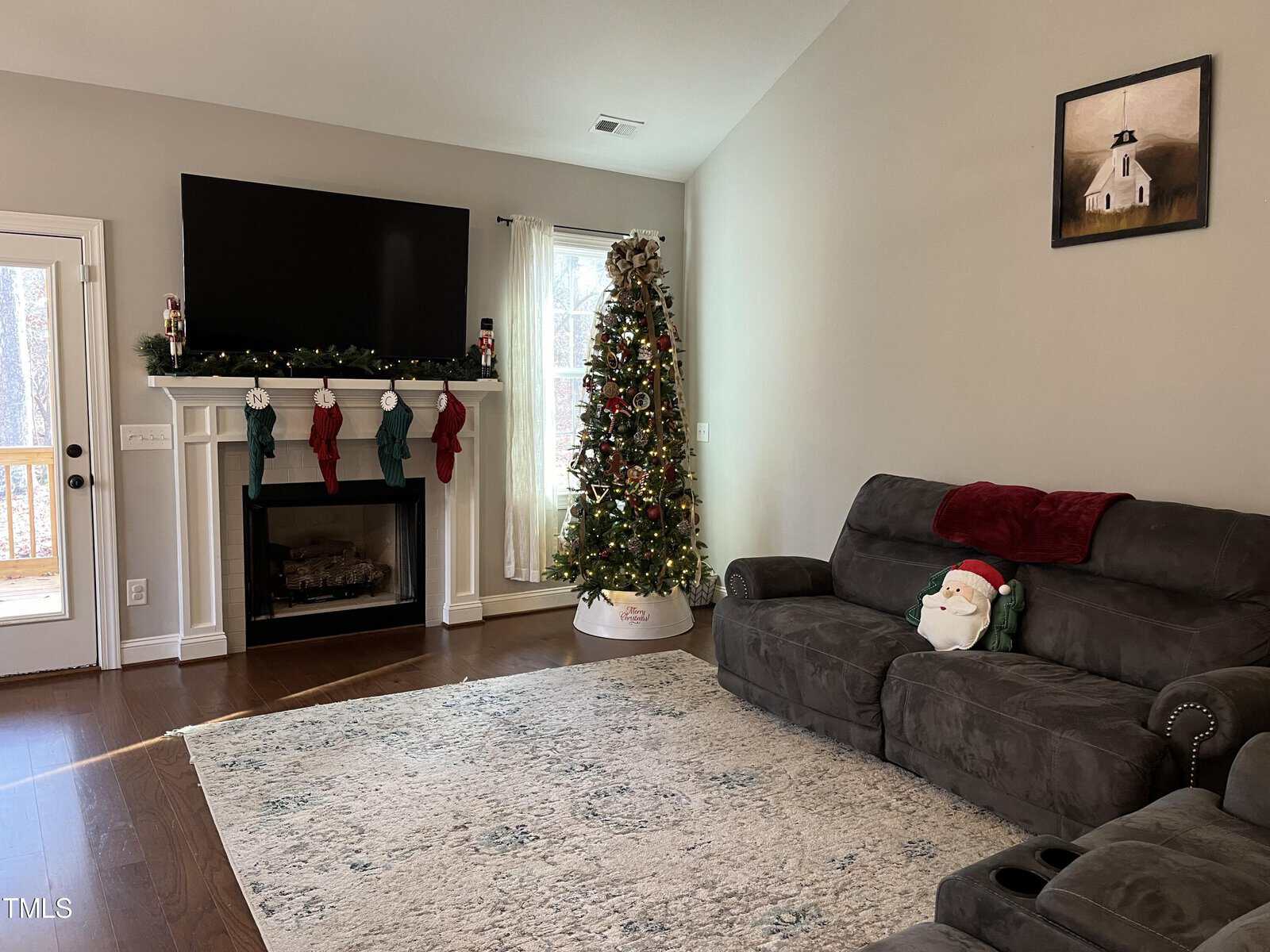 A cozy living room with a large decorated Christmas tree near a window. A fireplace is adorned with garland and stockings, topped by a flat-screen TV. A plush gray sofa features a Santa-themed pillow. Sunlight streams in through a door on the left.