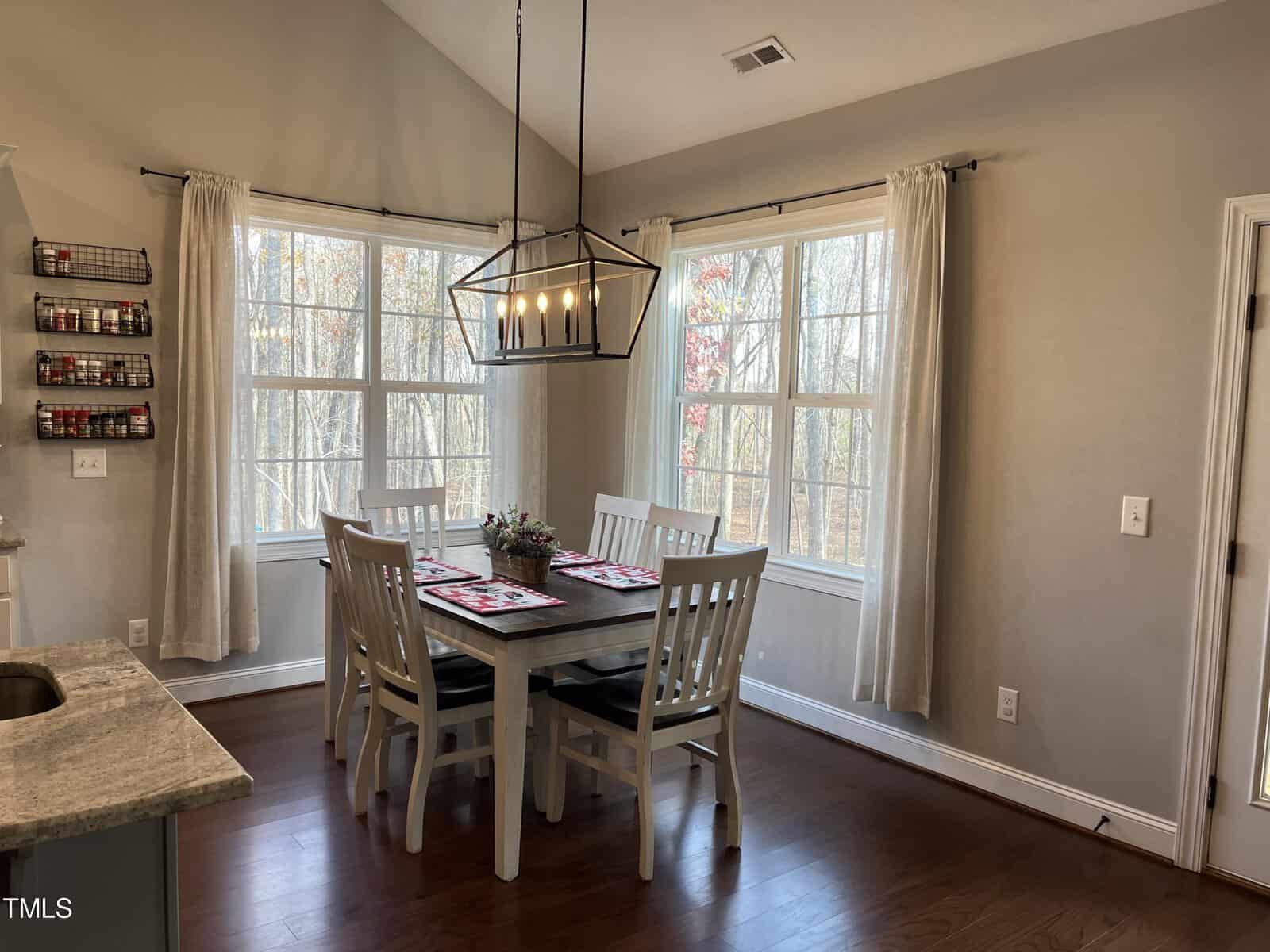 Dining area with a wooden table set for four and white chairs. A modern chandelier hangs above. Large windows provide natural light and a view of trees outside. Neutral colors and hardwood floors create a cozy atmosphere.
