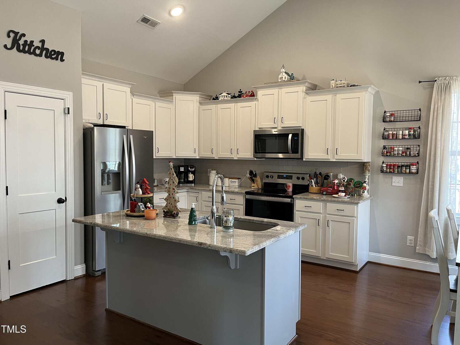 A modern kitchen with white cabinets, stainless steel appliances, and a granite island. The room is decorated with small household ornaments, and a sign reading "Kitchen" is above the doorway. Natural light fills the space through the window.