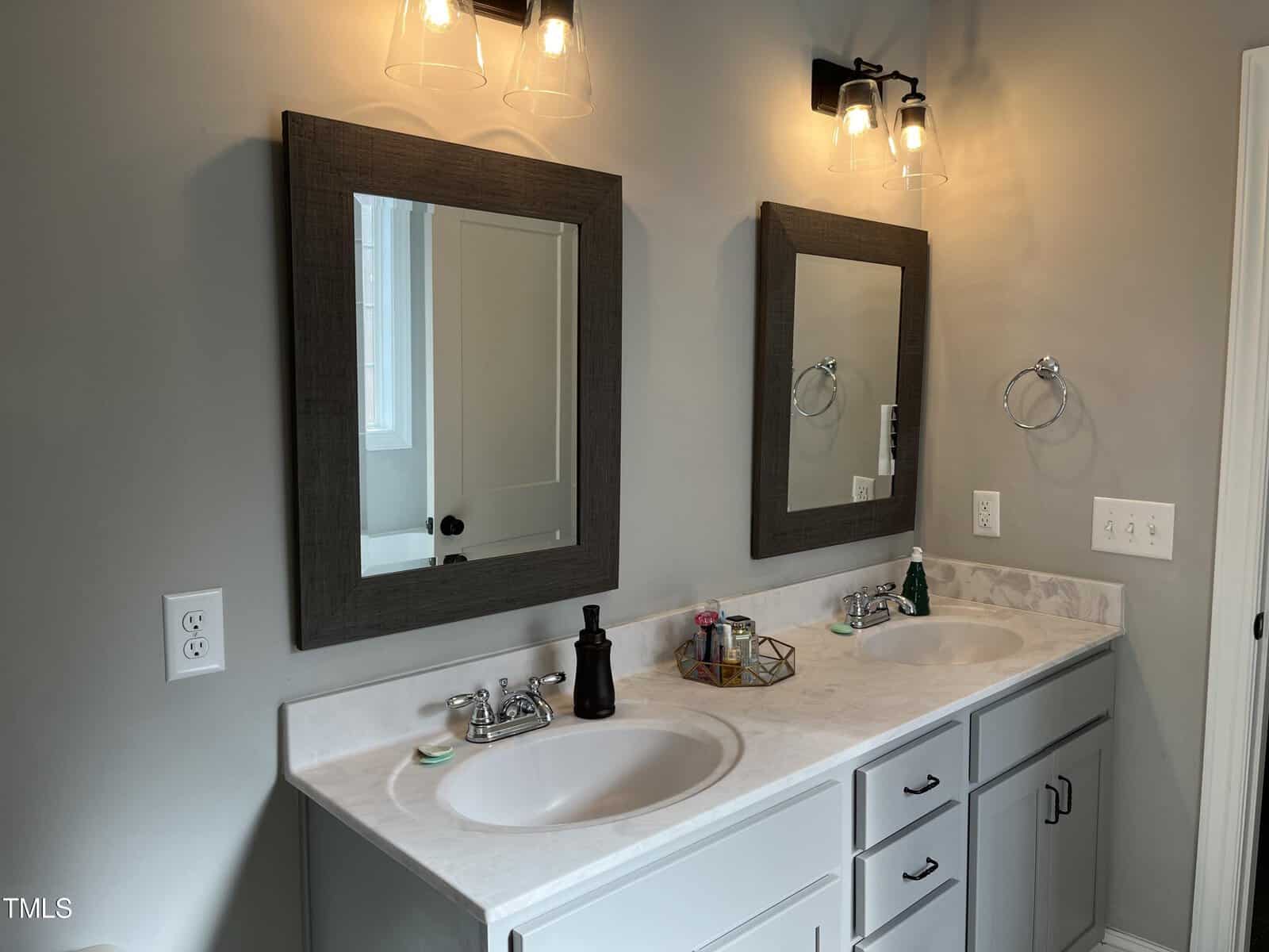 A modern bathroom features a double vanity with two rectangular mirrors. Each sink has a soap dispenser and toiletries. Two wall-mounted light fixtures illuminate the area. The countertop is marble, and the cabinets are pale gray.