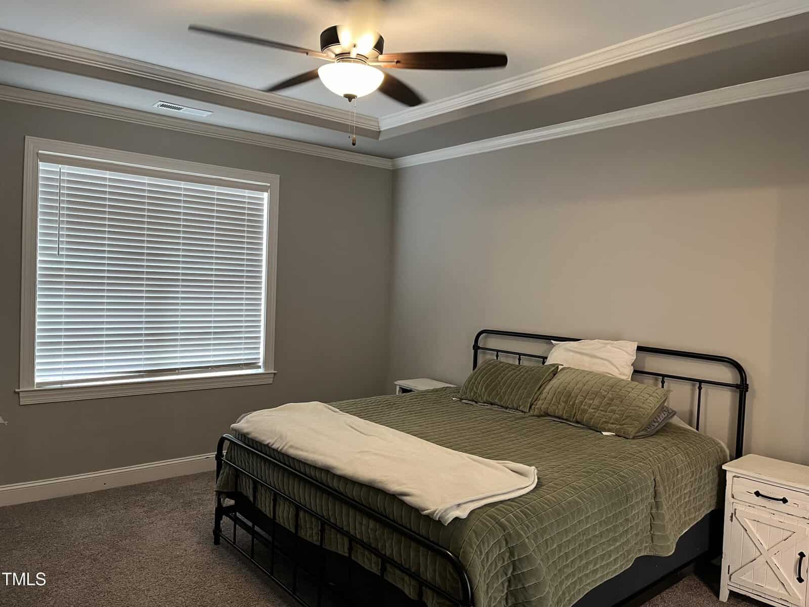 A bedroom with a large bed covered in a green quilt and white pillows. There's a ceiling fan with lights above, a window with closed blinds, and two white nightstands flanking the bed. The walls are painted light gray.