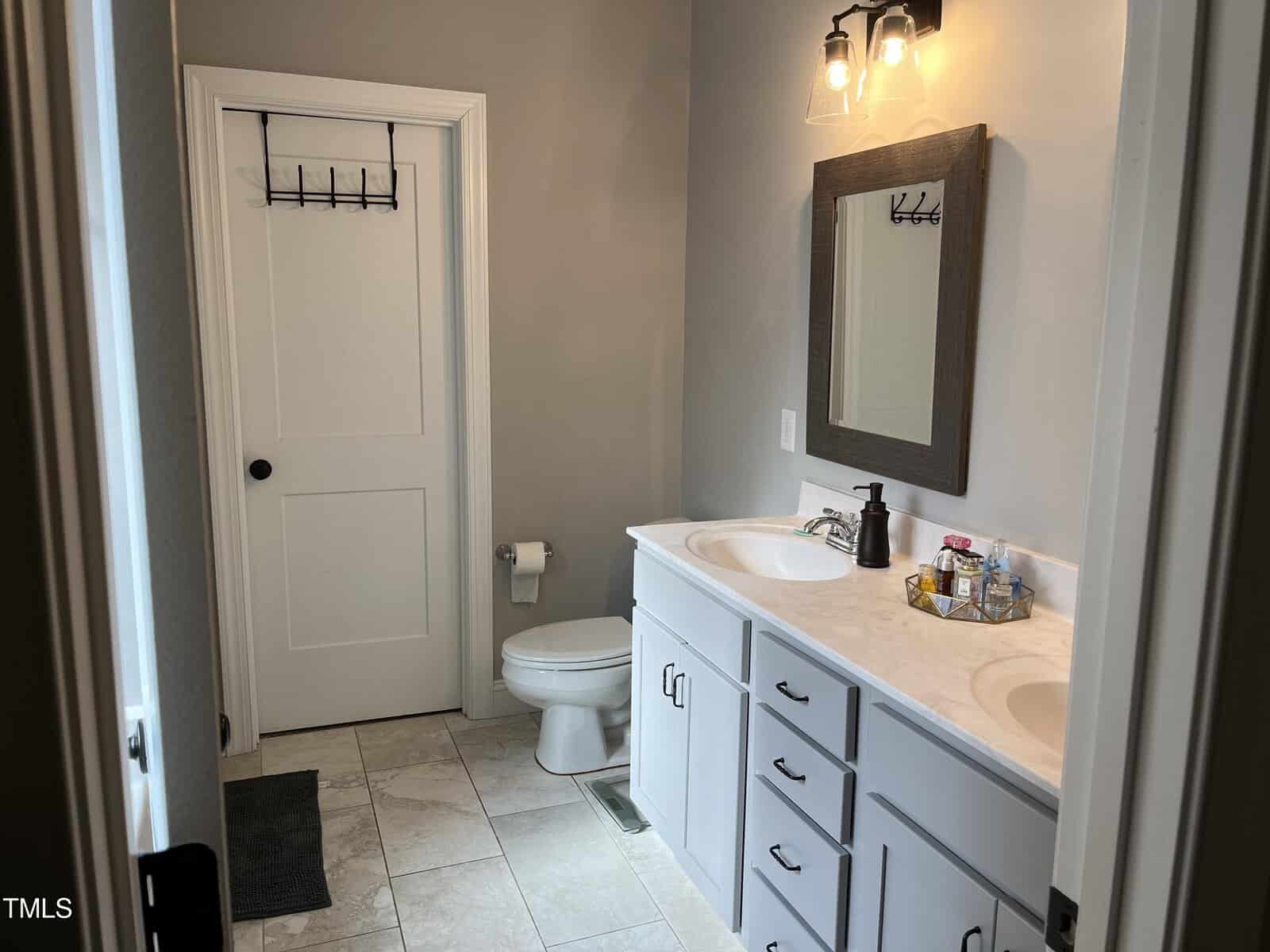 A bathroom with a double sink vanity, white cabinets, and a large mirror. The room has gray walls, tile flooring, and features a closed door with a towel hook and a toilet next to the sinks. Lighting fixtures are mounted above the mirror.