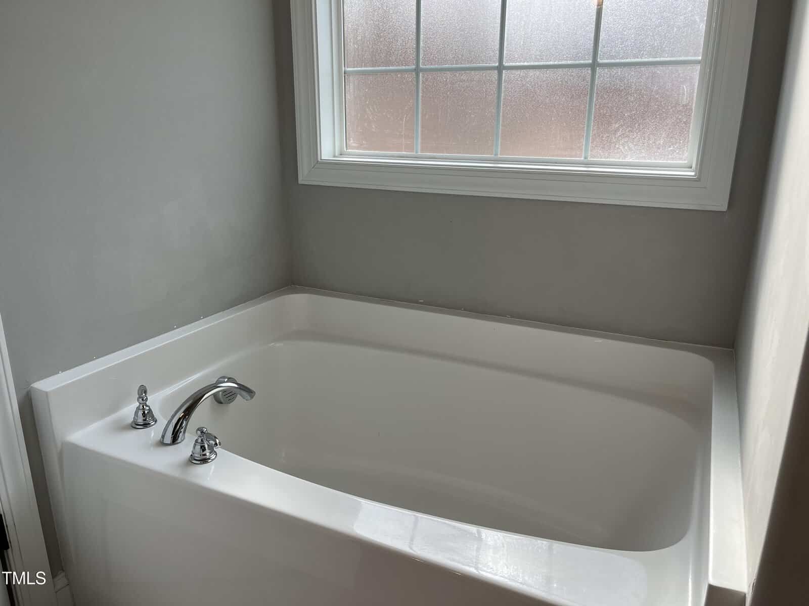 A clean, white bathtub with chrome faucet fixtures is situated below a large frosted window in a bathroom with light gray walls. Natural light is softly illuminating the space.