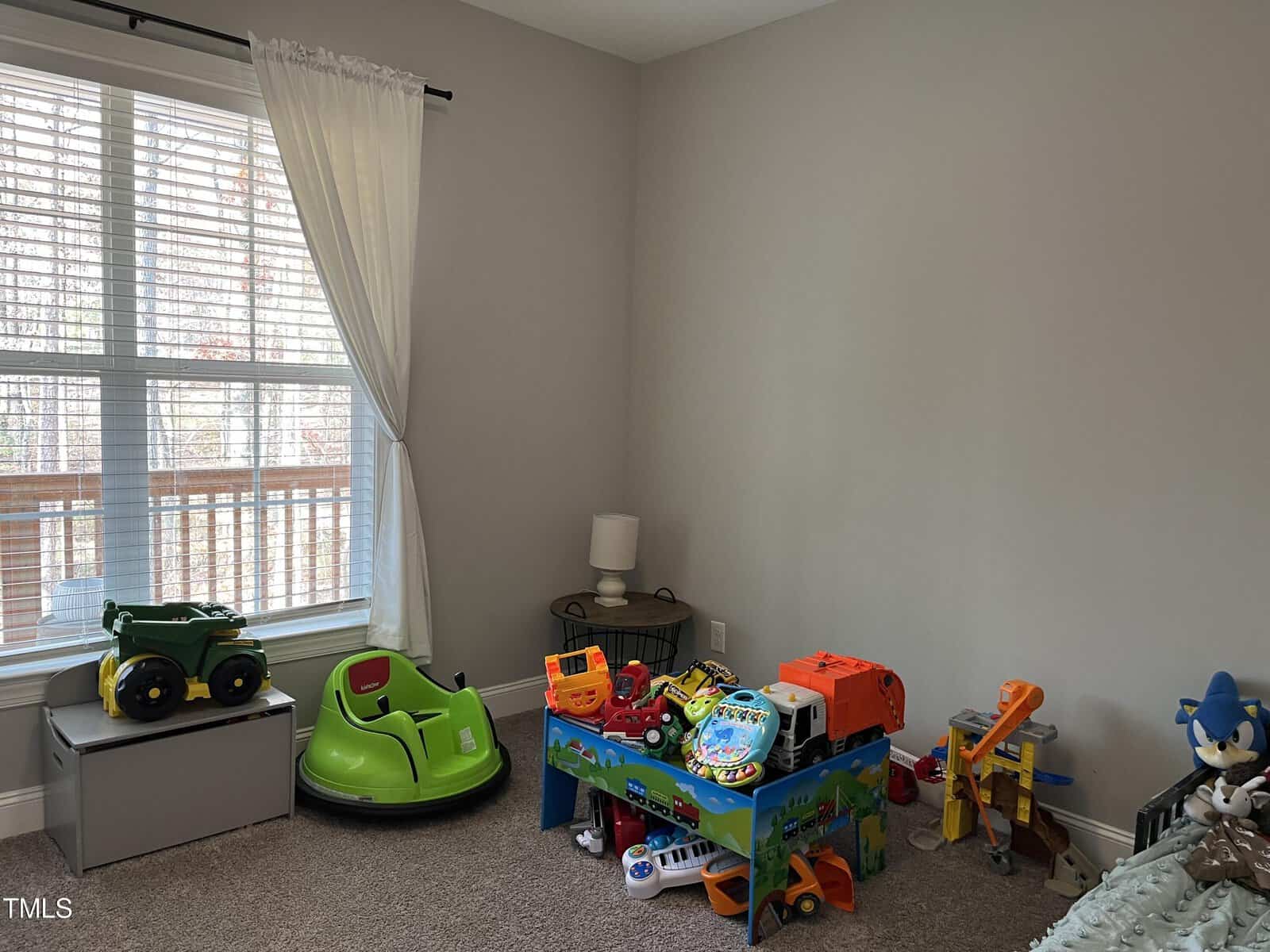 A playroom with a variety of toys, including a green toy car, trucks, and building blocks. A window with white curtains allows natural light in. A storage box and a small nightstand with a lamp are also visible in the room.