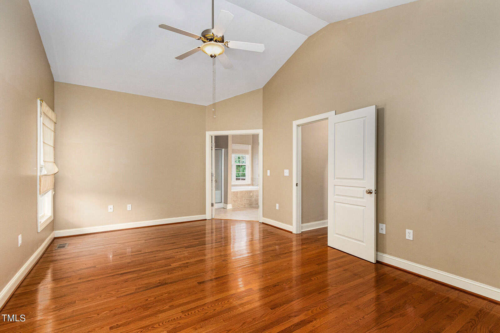 An empty room with light brown walls and polished wooden floors. There's a ceiling fan above, two windows on the left, and an open door leading to a bathroom. The room is well-lit with natural light.