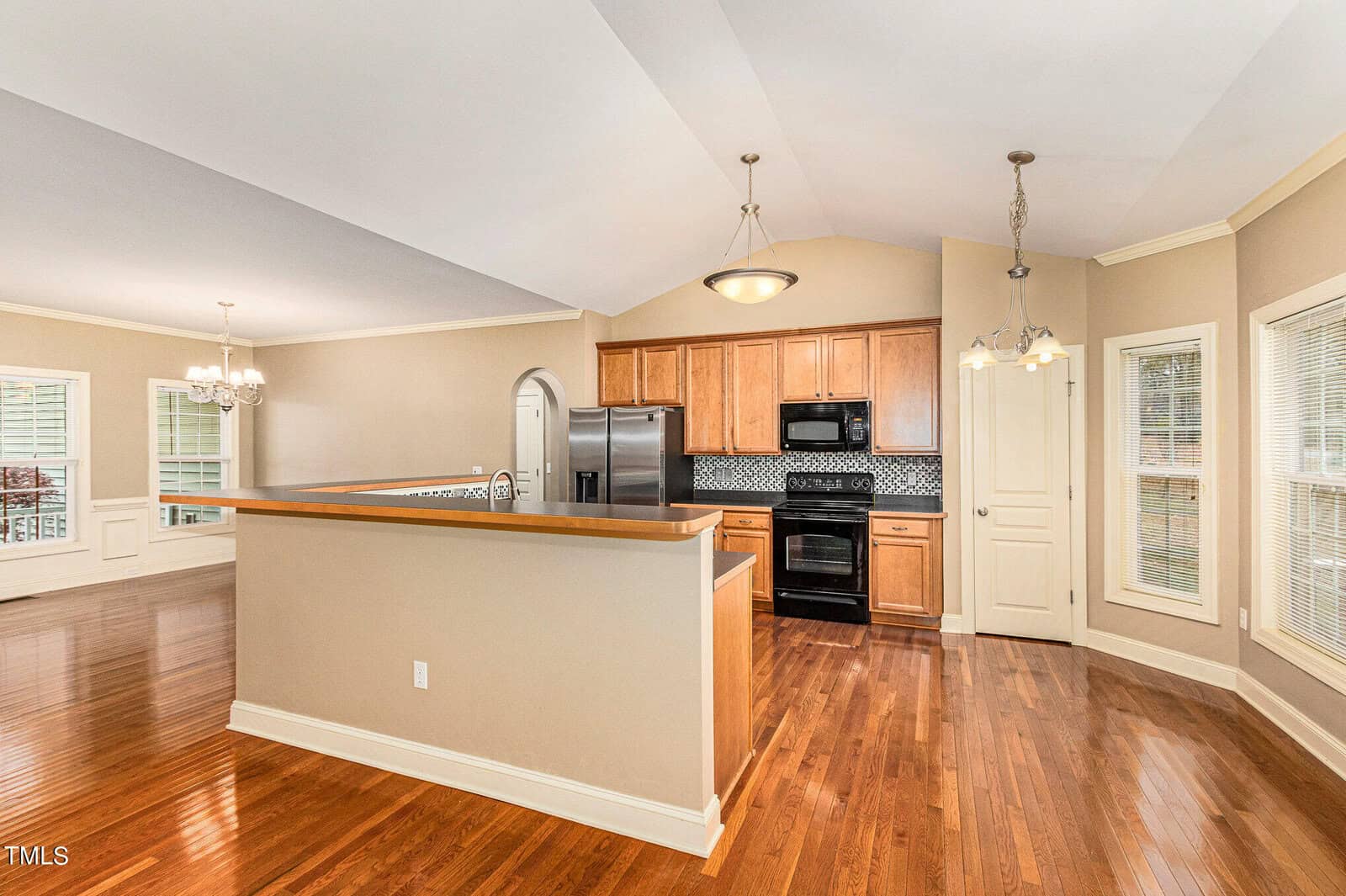 A modern kitchen with light wooden cabinets, black appliances, and a central island with a wooden countertop. The kitchen is adjacent to an open-plan dining area with large windows and hardwood floors. Two pendant lights hang above the island.