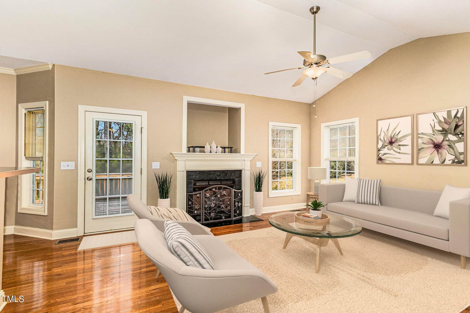 A spacious living room with high ceilings features a modern fireplace, tan walls, and wooden floors. Two beige sofas face a glass coffee table, and large windows allow natural light to fill the room. A ceiling fan is centered above.