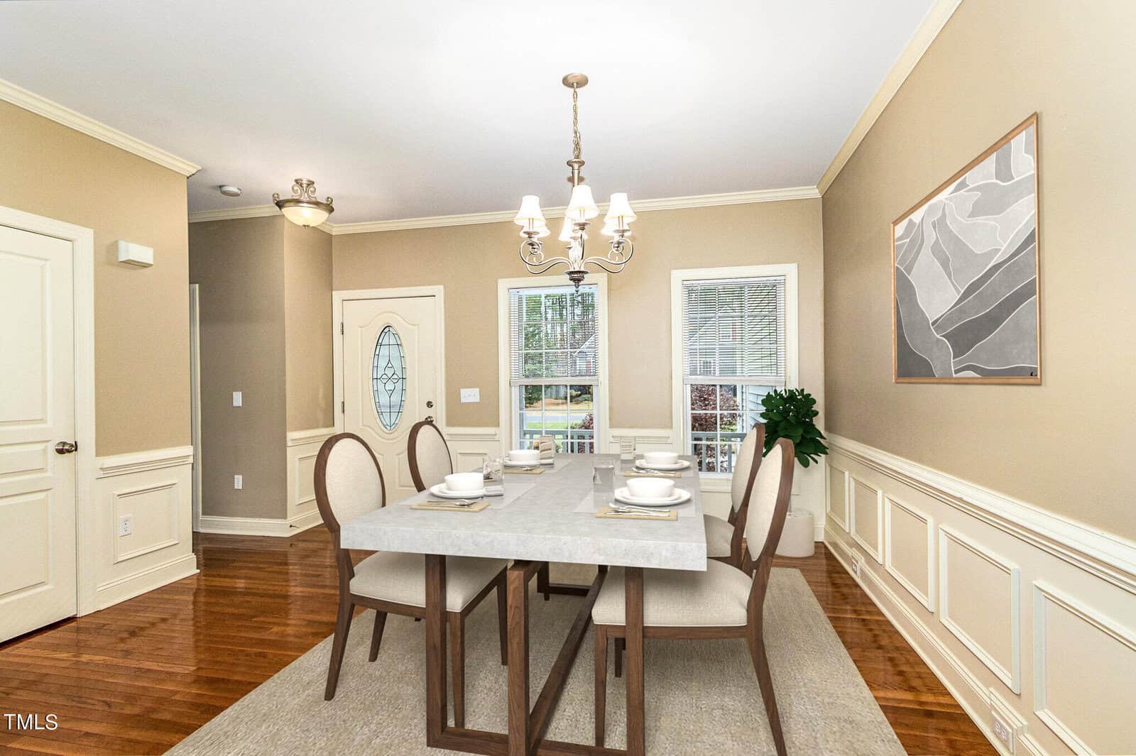 A dining room with a large rectangular table set for six, featuring cream-colored plates and glasses. The room has beige walls, wood flooring, a chandelier, a painting on the wall, and windows with white trim, letting in natural light.