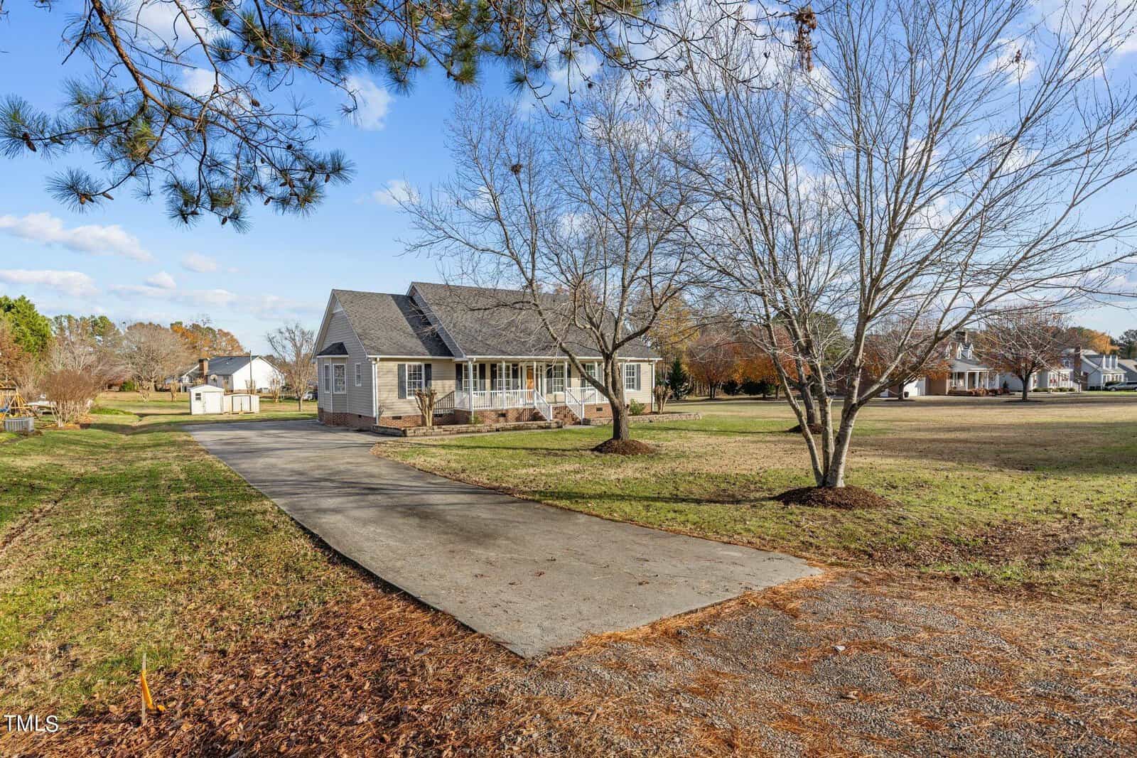 A quiet residential scene featuring a house with a gray roof and a sunroom extension, located at the end of a long driveway. The yard is spacious with bare trees and surrounded by other homes, under a partly cloudy sky.