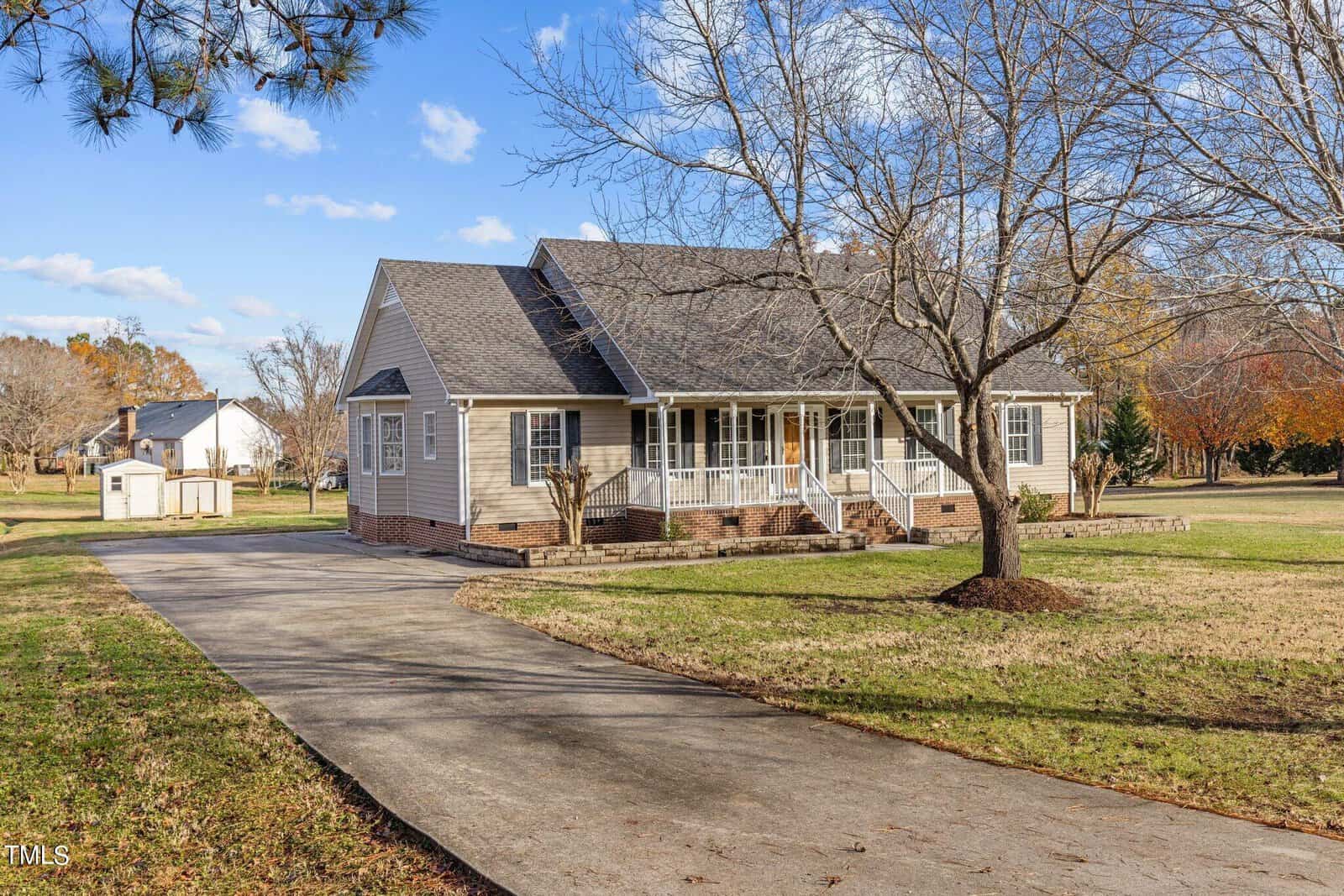 A single-story beige house with a porch sits on a large, grassy lawn. The driveway curves to the left, leading to the front porch. Trees are scattered throughout the yard, and the sky is clear with a few clouds.