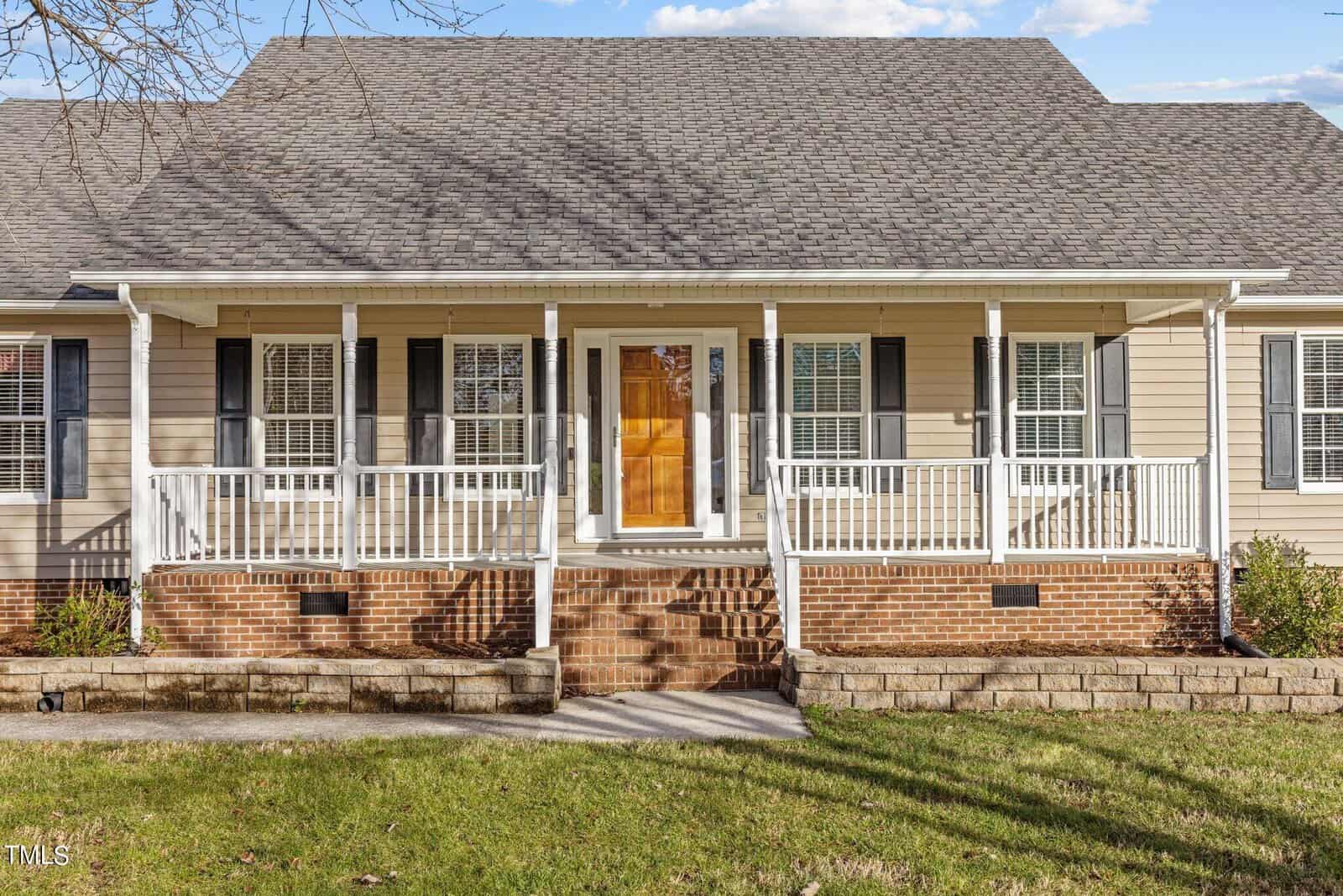 A beige house with a gabled roof and a brick foundation. It features a wooden front door surrounded by glass panels, flanked by large windows with dark shutters. A white-railed porch runs the width of the house, accessible by brick steps.