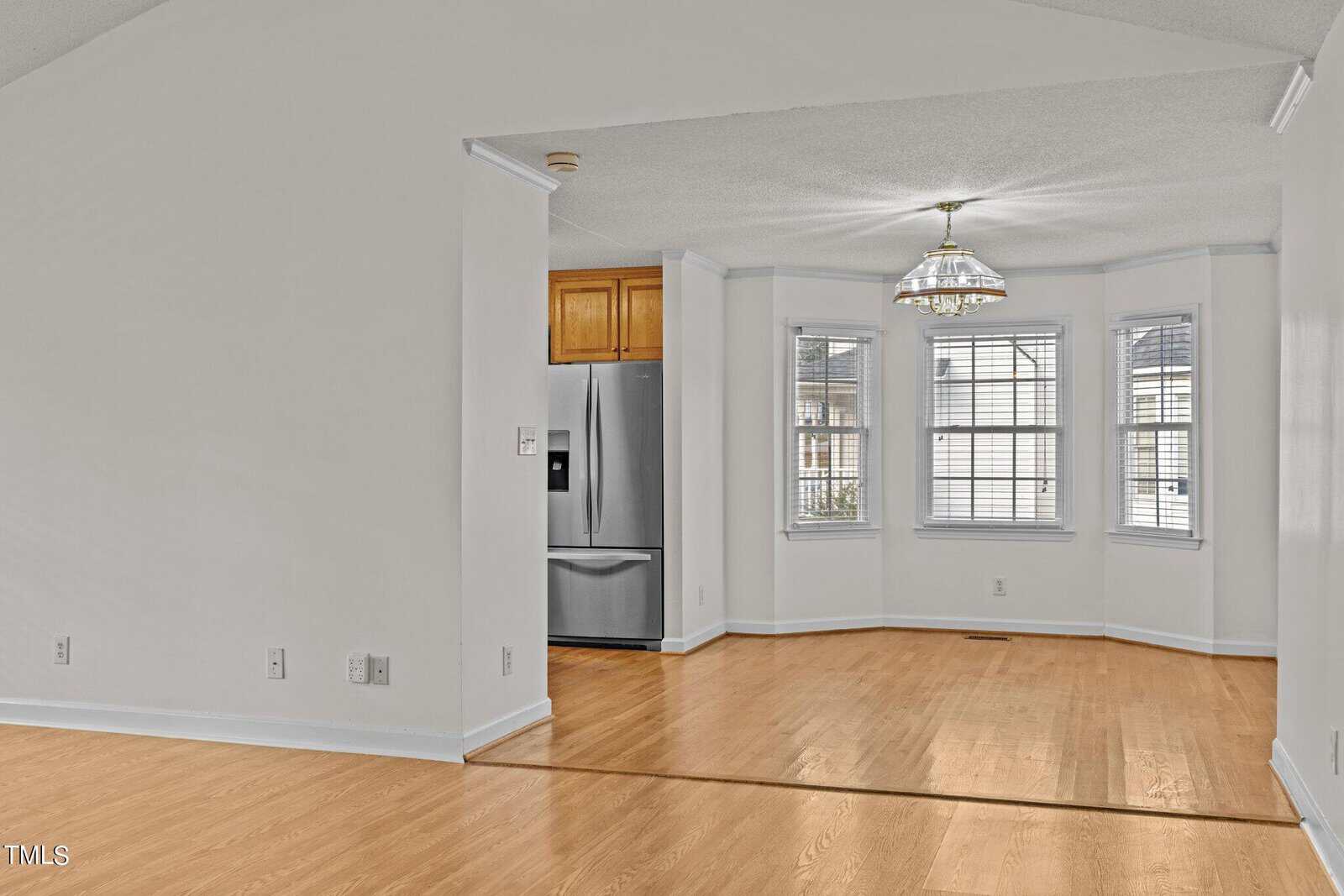 An empty room with light wood flooring and white walls. There's a bay window in the back with a multi-pane design and a hanging light fixture. A stainless steel refrigerator can be seen partially in an adjacent kitchen nook.