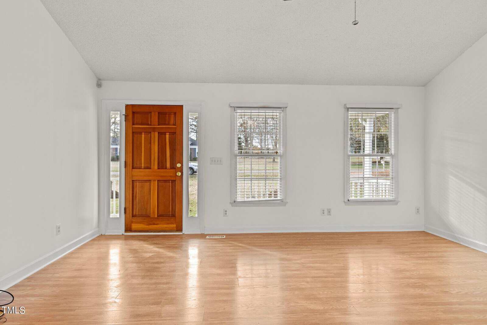A bright, empty room with light wood flooring, white walls, and a wooden front door. Two tall windows on the right let in natural light. The space has a simple, clean look.