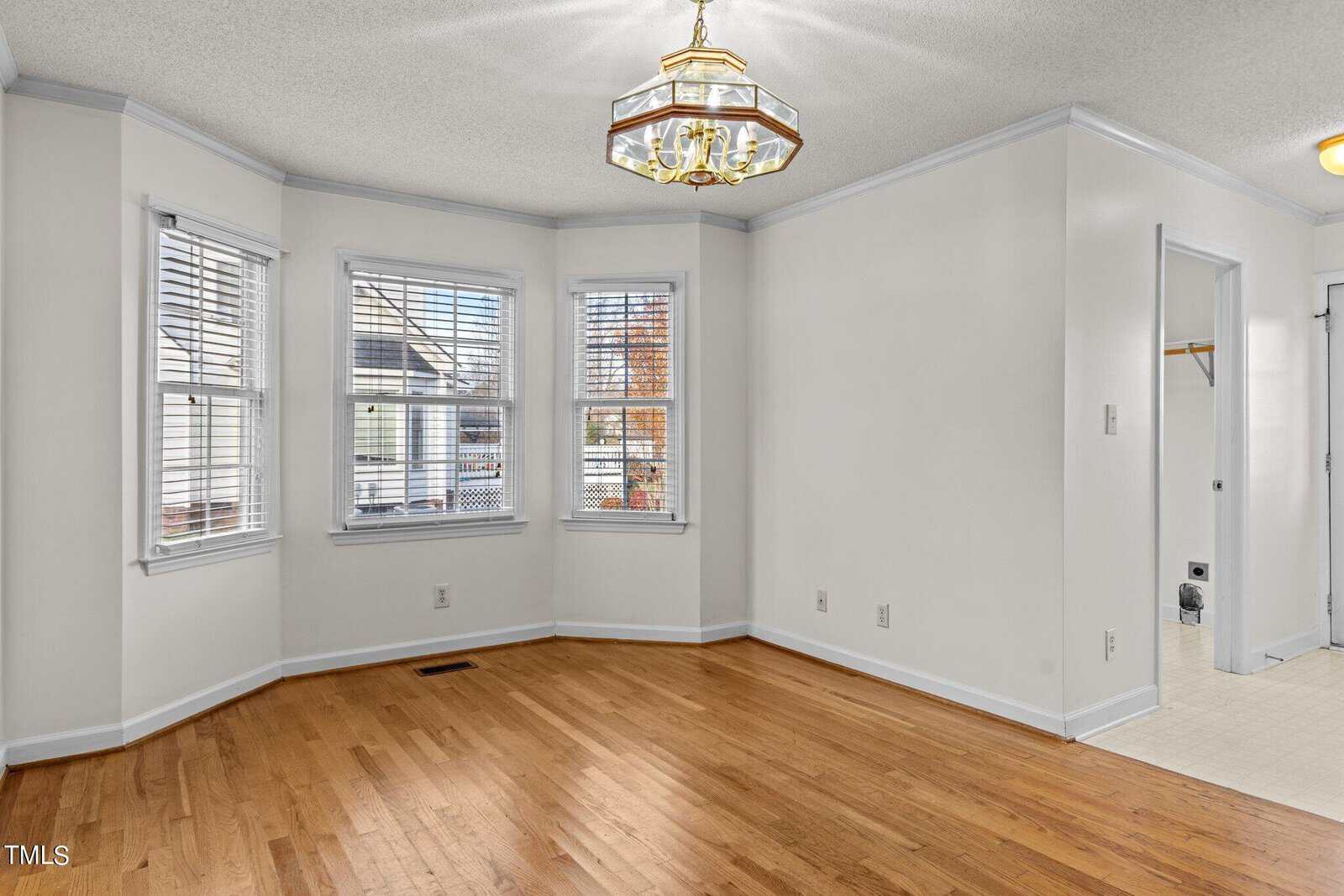 A bright room with wooden floors, cream walls, and a bay window featuring white blinds. A brass chandelier hangs from the ceiling. A small adjacent area with tiled flooring and an open door leading to another room is visible.