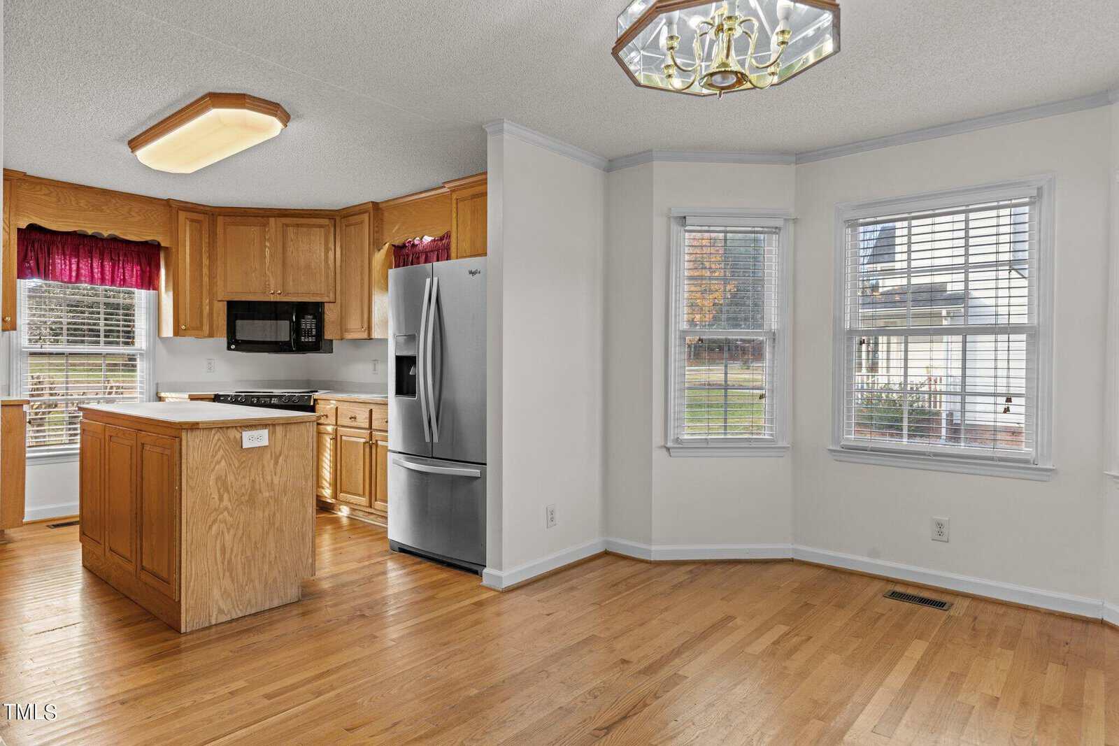 Bright kitchen with wooden cabinets, stainless steel appliances, and a central island. Adjoining dining area has large windows and hardwood floors. A hanging light fixture provides illumination.