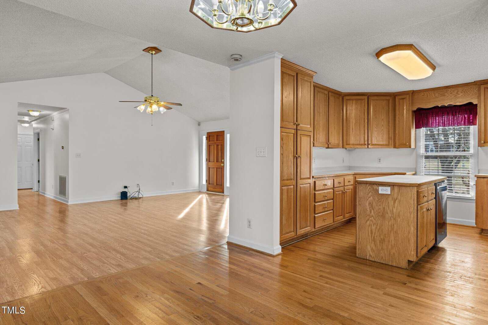 Open-concept living space with wooden flooring. The kitchen has wooden cabinets, a small island, and a dishwasher. A hanging light fixture is above the dining area, and the room has large windows with red valances, letting in natural light.