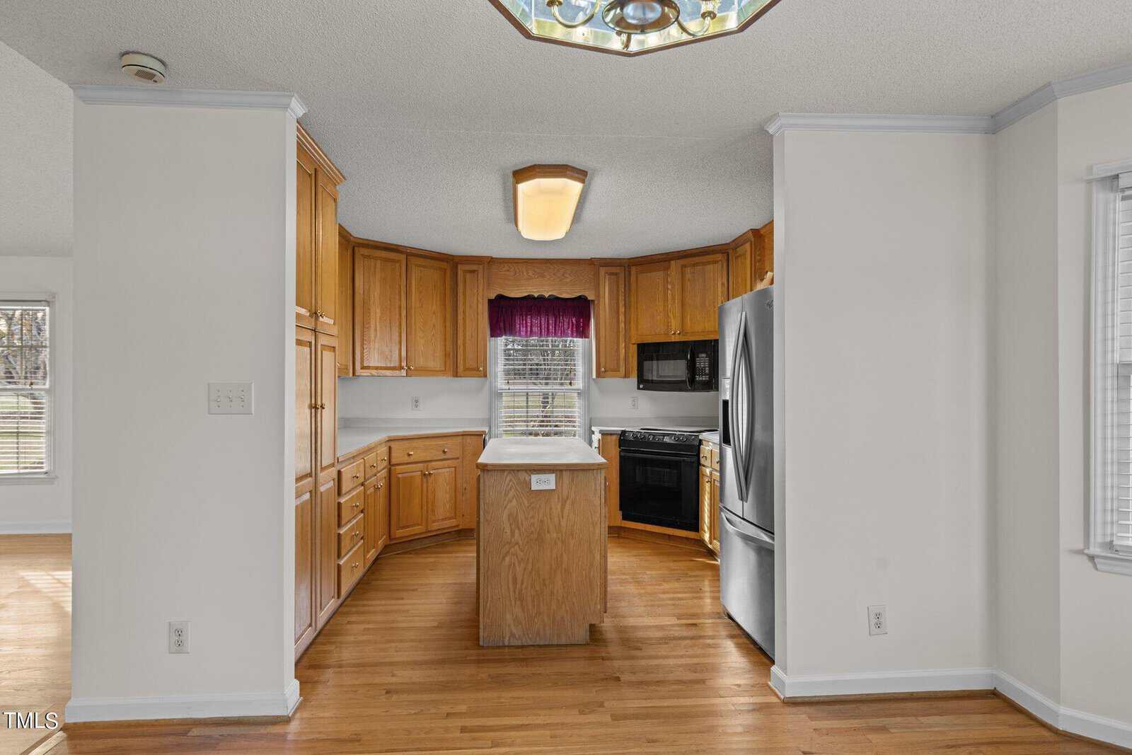 A kitchen with wooden cabinets, an island, beige countertops, a stainless steel refrigerator, and a black oven. A ceiling light fixture is above the island, and a purple valance decorates the window. Light wood flooring completes the look.