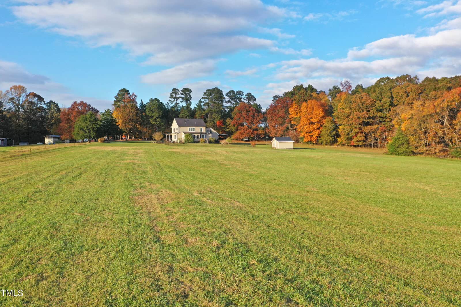 A large, open green field with a two-story house in the distance, surrounded by colorful autumn trees under a partly cloudy sky. A small shed is also visible near the house.