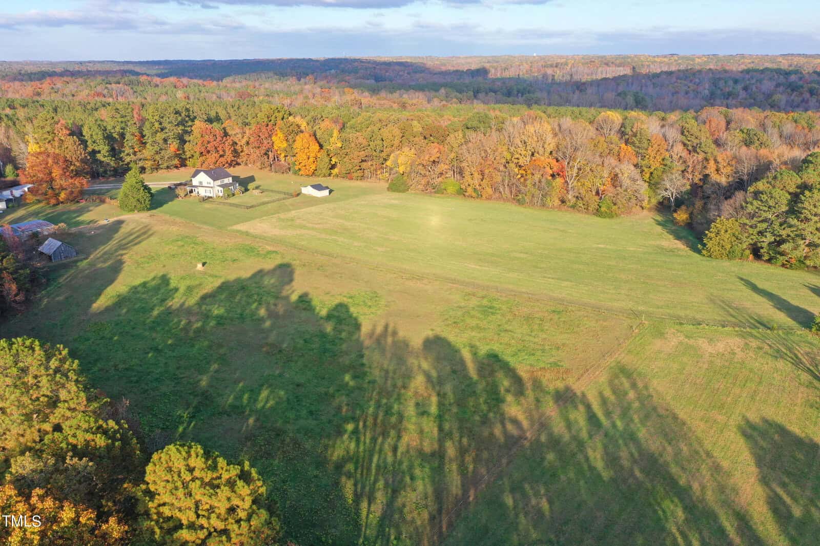 Aerial view of a rural landscape during autumn. A few houses are scattered among expansive green fields and vibrant trees with fall foliage. Shadows from the trees stretch across the ground under a partly cloudy sky.